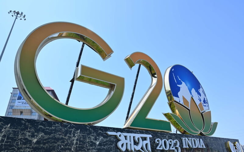 Ein gold umrandeter Schriftzug mit den Lettern "G20", der aus den Farben grün und orange besteht, steht neben dem Symbol einer Erde und einer Lotusblüte über dem Schriftzug "India 2023".