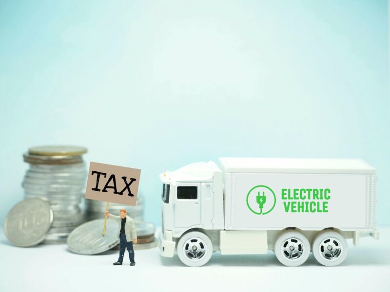The figure of a man with a sign that says Tax is in front of a toy truck that says Electric Vehicle.