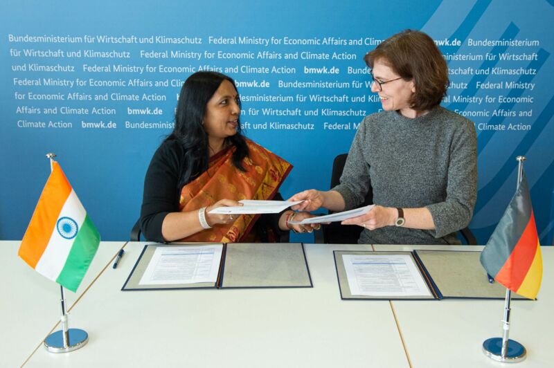 Two women facing each other exchange a document. In the foreground are a German and an Indian flag.