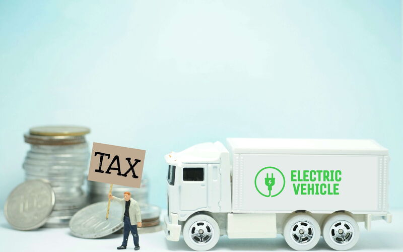 The figure of a man with a sign that says Tax is in front of a toy truck that says Electric Vehicle.