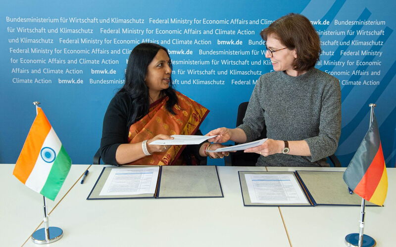 Zwei einander zugewandte Frauen tauschen ein Dokument aus. Im Vordergrund befinden sich eine deutsche und eine indische Flagge.