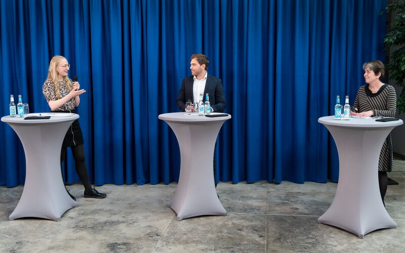 Christian Marian (DKE) and Maria Albert (DIN) discuss the topic "standardisation" together with the moderator from GPQI. They stand in front of a desk, in the background are blue curtains.